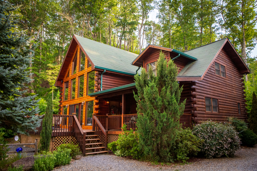 A log cabin with a front porch and lots of windows along the front sits in a heavily wooded area.