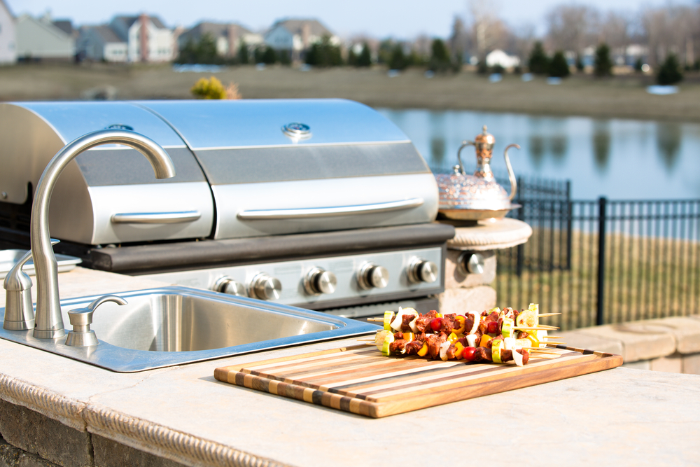 An outdoor kitchen with a built-in grill, sink, and a cutting board on the countertop with kebabs.