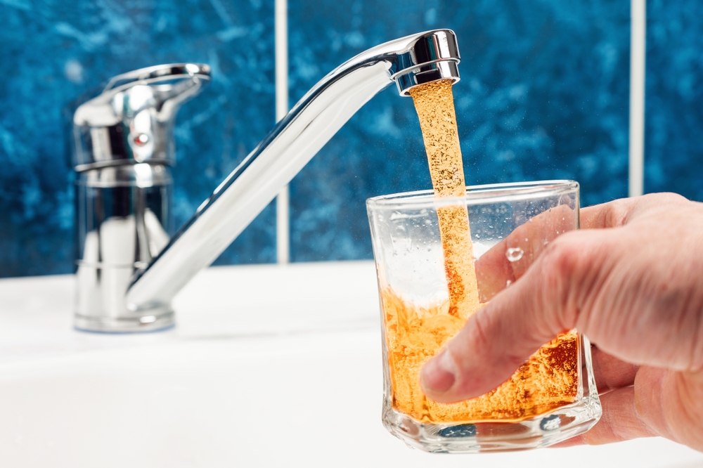 A person fills a glass from the tap with orange, rusty water.